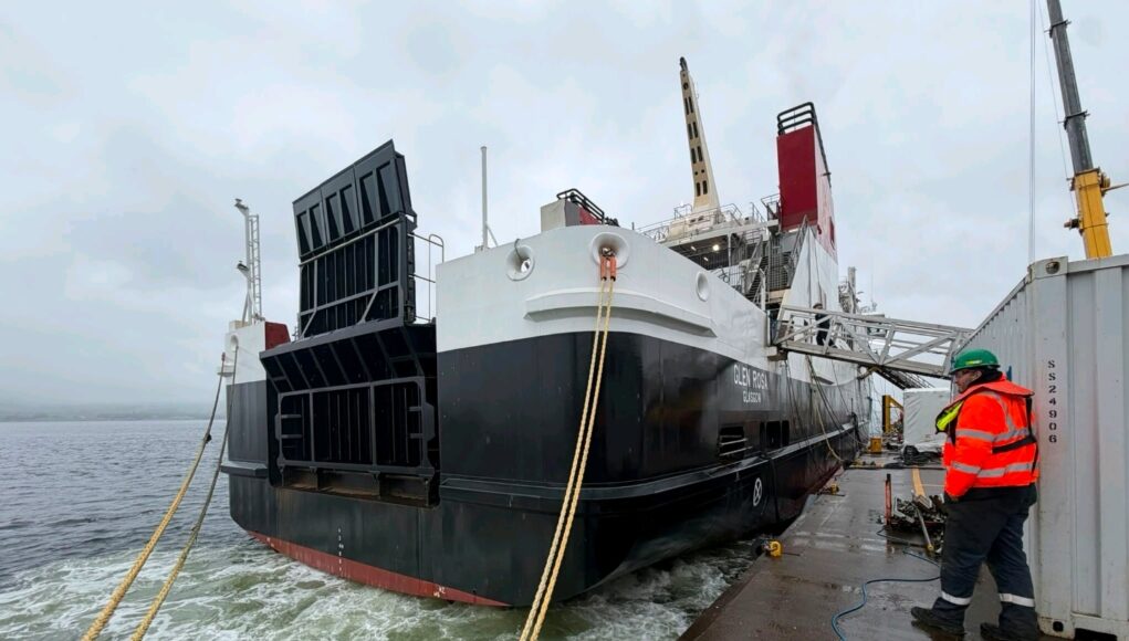 Large vessel docked at a quay with thick mooring lines; a worker in a high-visibility jacket and hard hat stands on the dock nearby.