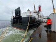 Glen Rosa’s engine roars to life at Ferguson Marine Large vessel docked at a quay with thick mooring lines; a worker in a high-visibility jacket and hard hat stands on the dock nearby.