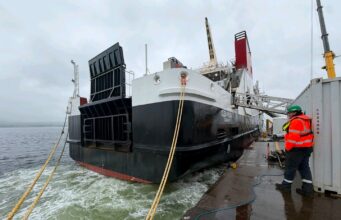 Glen Rosa’s engine roars to life at Ferguson Marine Large vessel docked at a quay with thick mooring lines; a worker in a high-visibility jacket and hard hat stands on the dock nearby.