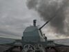 British warship opens fire with main gun Warship gun turret with a long cannon firing, dark smoke plume over the sea ahead of a gray, cloudy sky; ocean horizon in the distance.