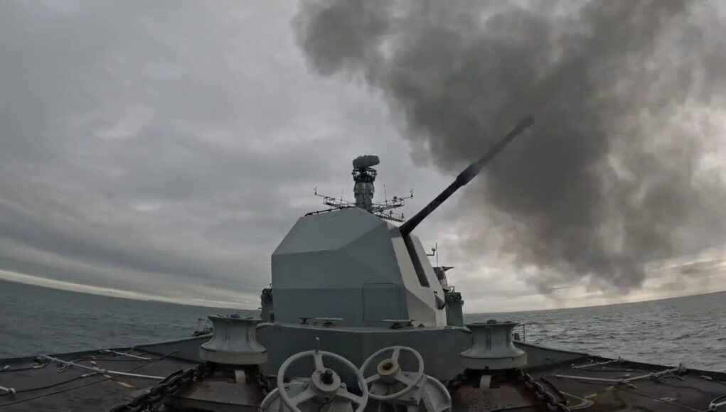 Warship gun turret with a long cannon firing, dark smoke plume over the sea ahead of a gray, cloudy sky; ocean horizon in the distance.