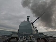 British warship opens fire with main gun Warship gun turret with a long cannon firing, dark smoke plume over the sea ahead of a gray, cloudy sky; ocean horizon in the distance.