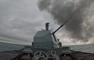 British warship opens fire with main gun Warship gun turret with a long cannon firing, dark smoke plume over the sea ahead of a gray, cloudy sky; ocean horizon in the distance.