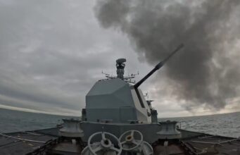 British warship opens fire with main gun Warship gun turret with a long cannon firing, dark smoke plume over the sea ahead of a gray, cloudy sky; ocean horizon in the distance.