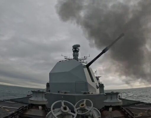 British warship opens fire with main gun Warship gun turret with a long cannon firing, dark smoke plume over the sea ahead of a gray, cloudy sky; ocean horizon in the distance.