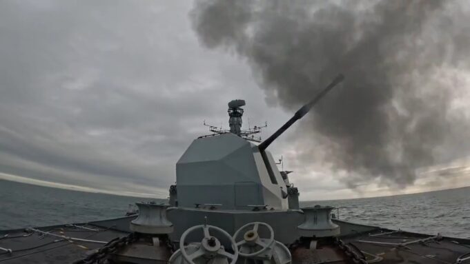 British warship opens fire with main gun Warship gun turret with a long cannon firing, dark smoke plume over the sea ahead of a gray, cloudy sky; ocean horizon in the distance.