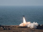 Small airplane launches a rocket from a coastal test site, flame and smoke plume rising toward the sky over the ocean.