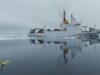 NATO turns drone Task Force X to the Arctic White research vessel sailing through icy water, its reflection visible; a small yellow unmanned aircraft floats in the foreground, mirrored on the surface.