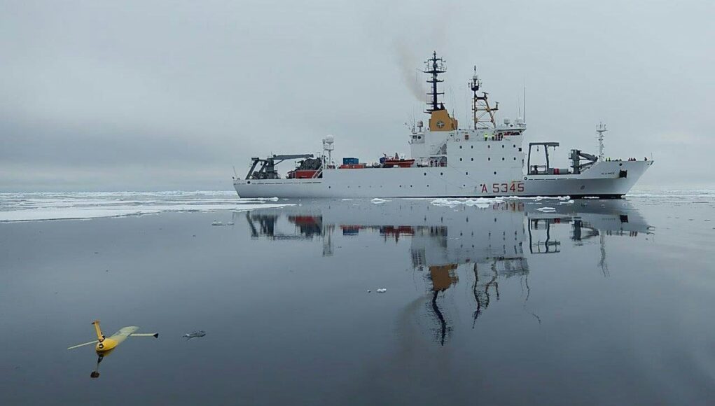 White research vessel sailing through icy water, its reflection visible; a small yellow unmanned aircraft floats in the foreground, mirrored on the surface.