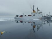 NATO turns drone Task Force X to the Arctic White research vessel sailing through icy water, its reflection visible; a small yellow unmanned aircraft floats in the foreground, mirrored on the surface.
