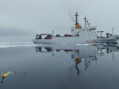 NATO turns drone Task Force X to the Arctic White research vessel sailing through icy water, its reflection visible; a small yellow unmanned aircraft floats in the foreground, mirrored on the surface.