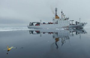NATO turns drone Task Force X to the Arctic White research vessel sailing through icy water, its reflection visible; a small yellow unmanned aircraft floats in the foreground, mirrored on the surface.