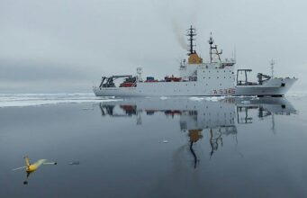 NATO turns drone Task Force X to the Arctic White research vessel sailing through icy water, its reflection visible; a small yellow unmanned aircraft floats in the foreground, mirrored on the surface.