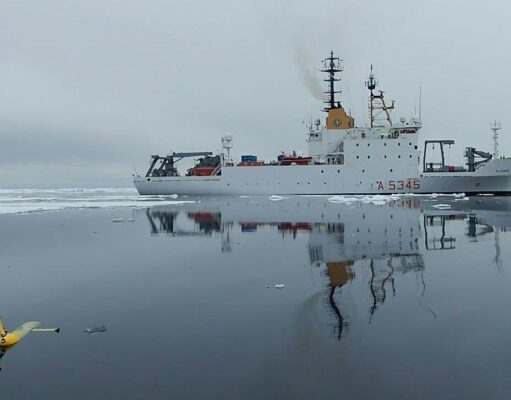 NATO turns drone Task Force X to the Arctic White research vessel sailing through icy water, its reflection visible; a small yellow unmanned aircraft floats in the foreground, mirrored on the surface.