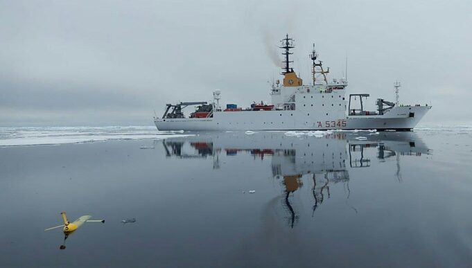 NATO turns drone Task Force X to the Arctic White research vessel sailing through icy water, its reflection visible; a small yellow unmanned aircraft floats in the foreground, mirrored on the surface.