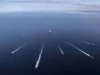 Aerial view of several ships at sea, each leaving white wake trails on the blue ocean against a distant horizon centralizing a larger vessel nearby.