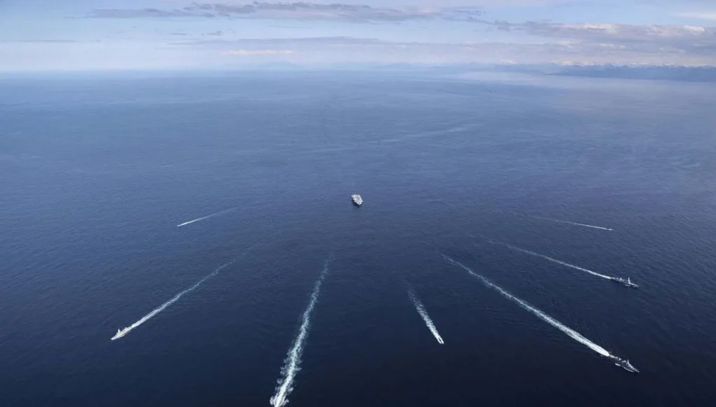 The Dynamic Mongoose participants spread out from HMS Prince of Wales Aerial view of several ships at sea, each leaving white wake trails on the blue ocean against a distant horizon centralizing a larger vessel nearby.