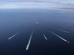 British aircraft carrier joins sub-hunt off Norway Aerial view of several ships at sea, each leaving white wake trails on the blue ocean against a distant horizon centralizing a larger vessel nearby.