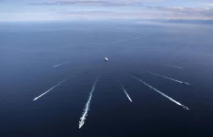 Aerial view of several ships at sea, each leaving white wake trails on the blue ocean against a distant horizon centralizing a larger vessel nearby.
