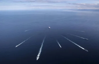 British aircraft carrier joins sub-hunt off Norway Aerial view of several ships at sea, each leaving white wake trails on the blue ocean against a distant horizon centralizing a larger vessel nearby.