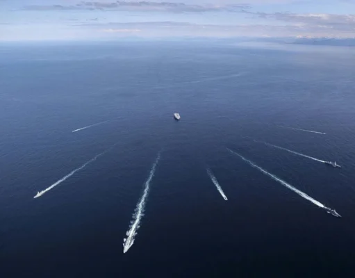 British aircraft carrier joins sub-hunt off Norway Aerial view of several ships at sea, each leaving white wake trails on the blue ocean against a distant horizon centralizing a larger vessel nearby.