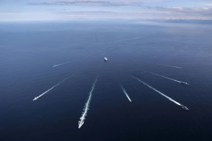 British aircraft carrier joins sub-hunt off Norway Aerial view of several ships at sea, each leaving white wake trails on the blue ocean against a distant horizon centralizing a larger vessel nearby.