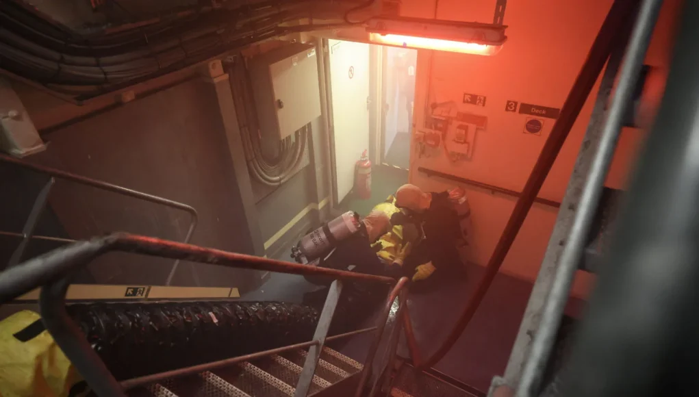 The firefighting party moves down one of the ships stairwells Firefighters in yellow turnout gear with breathing apparatus crouch in a smoke-filled ship corridor under red emergency lighting near a doorway.