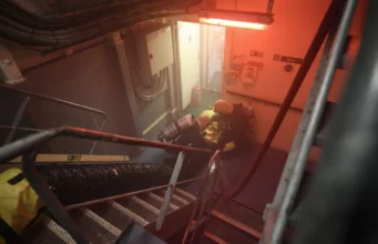 Firefighters in yellow turnout gear with breathing apparatus crouch in a smoke-filled ship corridor under red emergency lighting near a doorway.