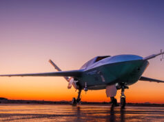 US Air Force orders uncrewed fighter into production Low-angle shot of a futuristic green aircraft parked on a wet runway at sunset, orange sky behind.
