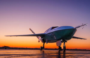 US Air Force orders uncrewed fighter into production Low-angle shot of a futuristic green aircraft parked on a wet runway at sunset, orange sky behind.