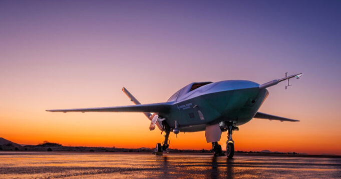 US Air Force orders uncrewed fighter into production Low-angle shot of a futuristic green aircraft parked on a wet runway at sunset, orange sky behind.