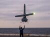 Romanian Navy official loses fingers to Shield AI V-BAT Person on a ship deck raises arms as a small helicopter hovers above, landing gear down, seawater on the horizon at dusk.