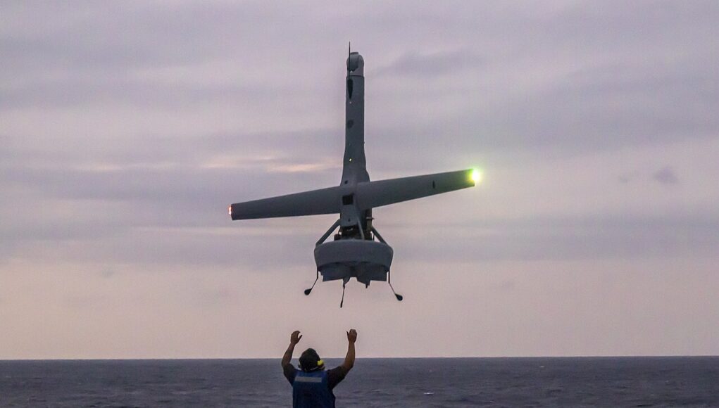 V-BAT_lands_on_USS_Green_Bay Person on a ship deck raises arms as a small helicopter hovers above, landing gear down, seawater on the horizon at dusk.