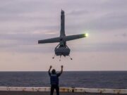 Romanian Navy official loses fingers to Shield AI V-BAT Person on a ship deck raises arms as a small helicopter hovers above, landing gear down, seawater on the horizon at dusk.