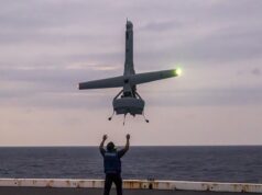 Person on a ship deck raises arms as a small helicopter hovers above, landing gear down, seawater on the horizon at dusk.