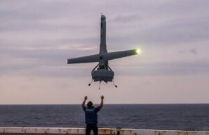 Person on a ship deck raises arms as a small helicopter hovers above, landing gear down, seawater on the horizon at dusk.