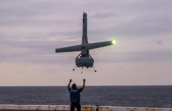 Romanian Navy official loses fingers to Shield AI V-BAT Person on a ship deck raises arms as a small helicopter hovers above, landing gear down, seawater on the horizon at dusk.