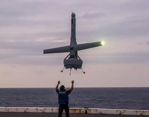 Romanian Navy official loses fingers to Shield AI V-BAT Person on a ship deck raises arms as a small helicopter hovers above, landing gear down, seawater on the horizon at dusk.