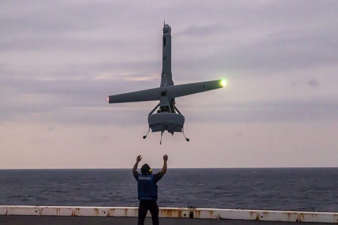 Person on a ship deck raises arms as a small helicopter hovers above, landing gear down, seawater on the horizon at dusk.