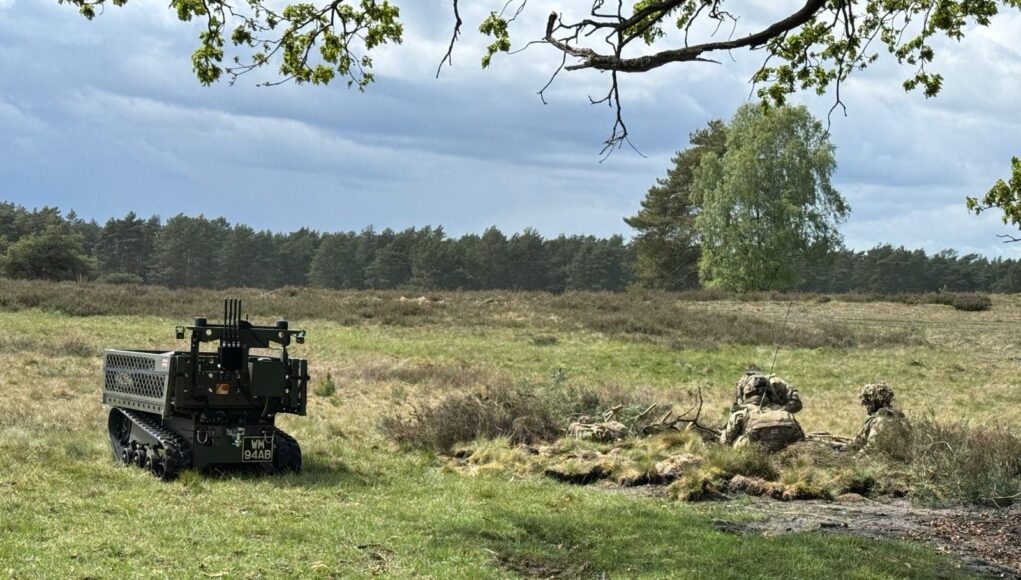 Autonomous unmanned ground vehicle on tracks in a grassy field, with soldiers in camouflage in a shallow trench to the right, under a cloudy sky.