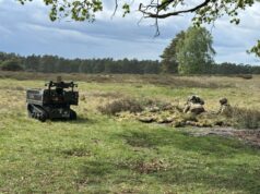 British Army picks DCE for first operational UGV fleet Autonomous unmanned ground vehicle on tracks in a grassy field, with soldiers in camouflage in a shallow trench to the right, under a cloudy sky.