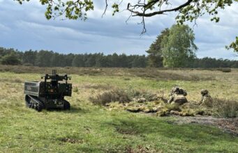 Autonomous unmanned ground vehicle on tracks in a grassy field, with soldiers in camouflage in a shallow trench to the right, under a cloudy sky.