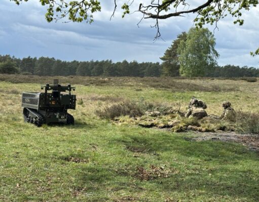 Autonomous unmanned ground vehicle on tracks in a grassy field, with soldiers in camouflage in a shallow trench to the right, under a cloudy sky.