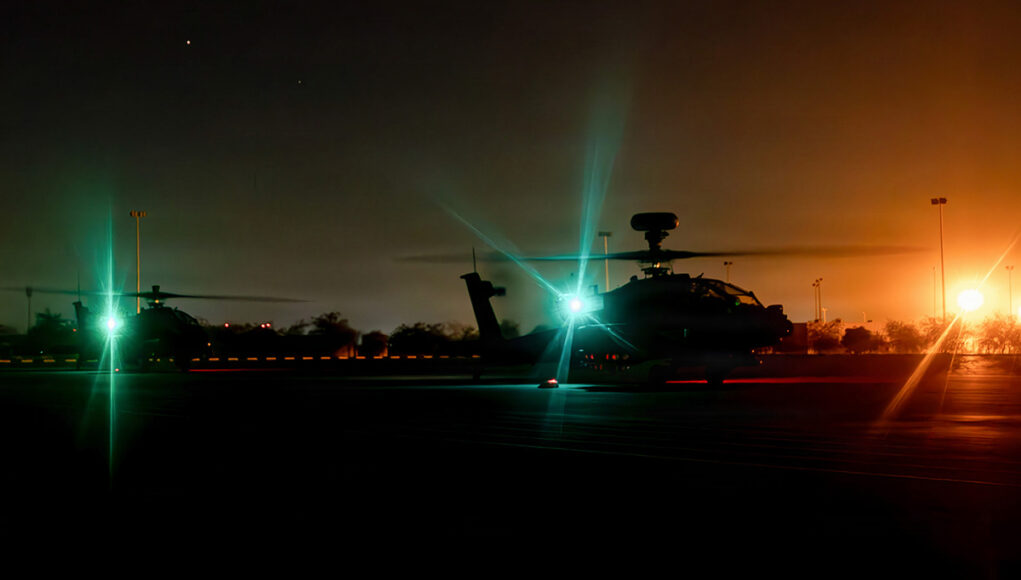 ah64e2 Nighttime scene of military helicopters on a runway with bright green and orange runway lights, silhouettes of rotors and equipment visible in the dark.