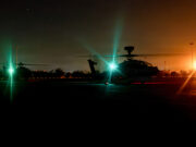 Nighttime scene of military helicopters on a runway with bright green and orange runway lights, silhouettes of rotors and equipment visible in the dark.