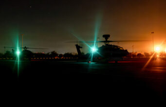 Nighttime scene of military helicopters on a runway with bright green and orange runway lights, silhouettes of rotors and equipment visible in the dark.