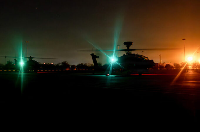 Nighttime scene of military helicopters on a runway with bright green and orange runway lights, silhouettes of rotors and equipment visible in the dark.