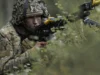 British troops test drone war tactics near Russian border Soldier in camouflage aiming a rifle while crouched among dense forest vegetation.