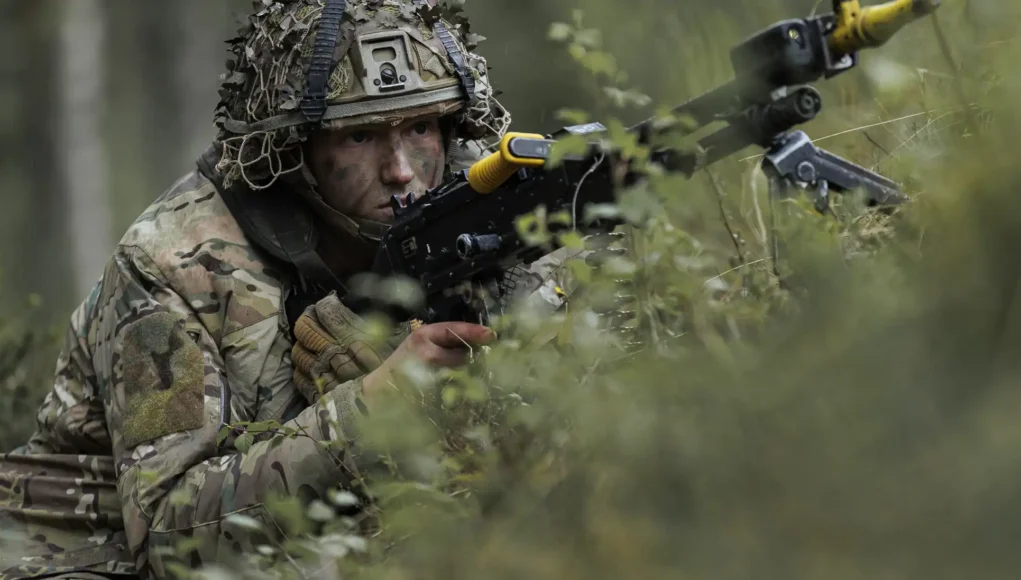 ahqcpl3-official-20260525-002 Soldier in camouflage aiming a rifle while crouched among dense forest vegetation.