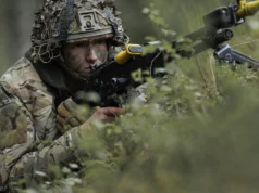 British troops test drone war tactics near Russian border Soldier in camouflage aiming a rifle while crouched among dense forest vegetation.